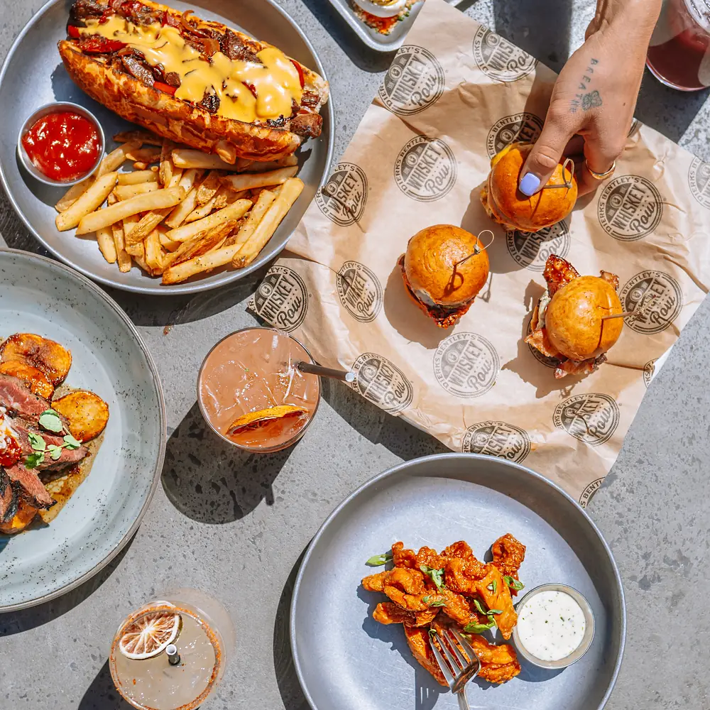 Overhead view of a Whiskey Row gastropub spread with a loaded hot dog and fries, sliders, wings and a cocktail